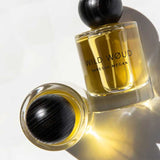 Two bottles of Vanessa Megan Wild Wøud 100% Natural Eau de Parfum 50ml, with golden liquid and black rounded caps, rest on a white surface—one upright and one lying down—casting soft shadows.