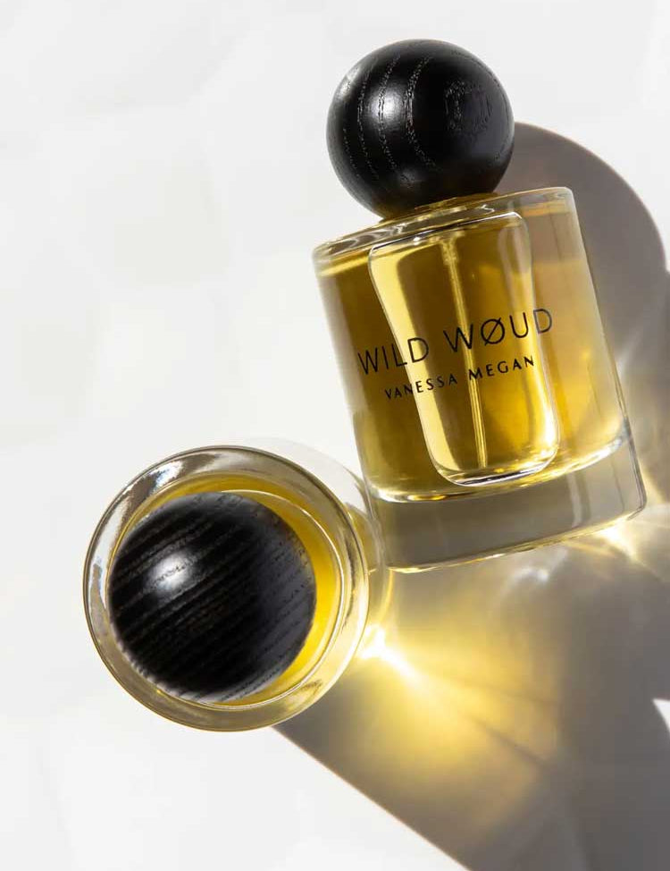 Two bottles of Vanessa Megan Wild Wøud 100% Natural Eau de Parfum 50ml, with golden liquid and black rounded caps, rest on a white surface, one upright and one lying down, casting soft shadows.