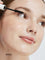 A close-up of a person with fair skin smiling slightly as they apply Ere Perez Avocado Waterproof Mascara 10ml to their upper lashes. The word Black is shown in the lower left corner.