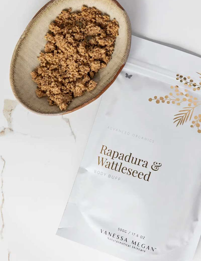 A beige bowl of brown, crumbly powder sits beside a white pouch labeled Vanessa Megan Rapadura & Wattleseed Body Buff 500g, an exfoliating and nourishing body polish, on a white marble surface.