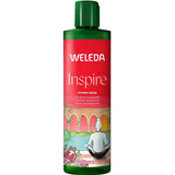 A green and red bottle of Weleda Inspire Pomegranate Shower Cream with a green cap, decorated with pomegranate illustrations and an image of a person by a pool with palm trees in the background.