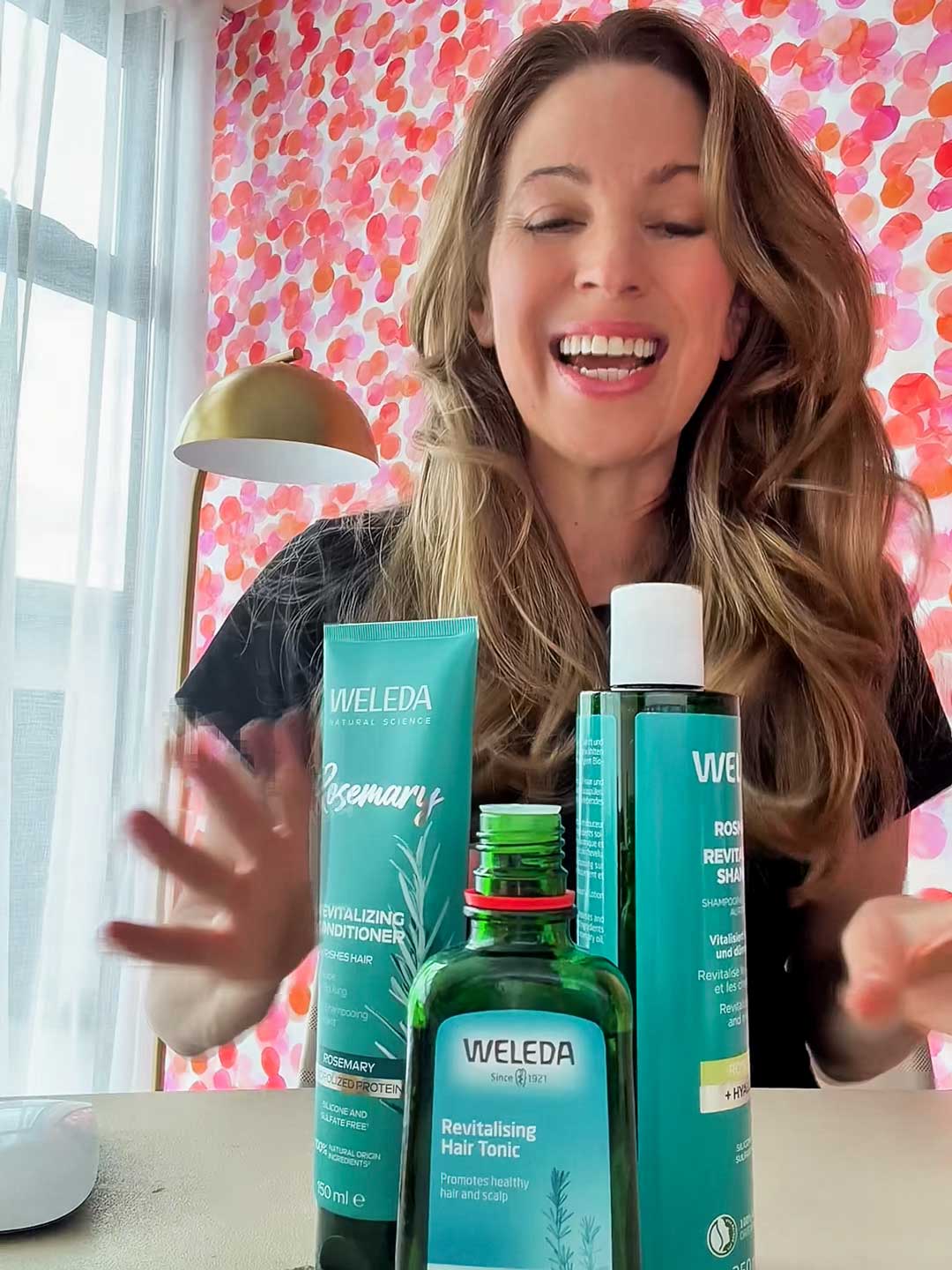 A smiling woman with long brown hair sits at a table with the Weleda Hair Bundle for Thin Hair Value Pack in front of her. A lamp and pink polka-dot wall brighten the cozy scene.