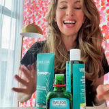 A smiling woman with long brown hair sits at a table with the Weleda Hair Bundle for Thin Hair Value Pack in front of her. A lamp and pink polka-dot wall brighten the cozy scene.