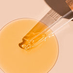 A glass dropper lies diagonally across a round pool of golden A bit Hippy Face Oil on a peach surface, casting a long shadow beside the glossy oil.
