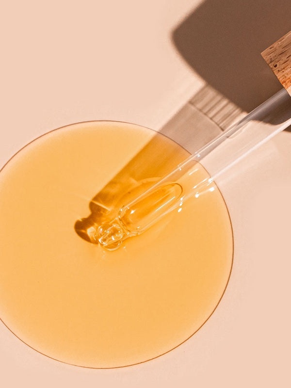 A glass dropper lies diagonally across a round pool of golden A bit Hippy Face Oil on a peach surface, casting a long shadow beside the glossy oil.