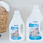 Two bottles of Abode Fabric Softener Zero on a white laundry bench beside a woven basket and folded towels