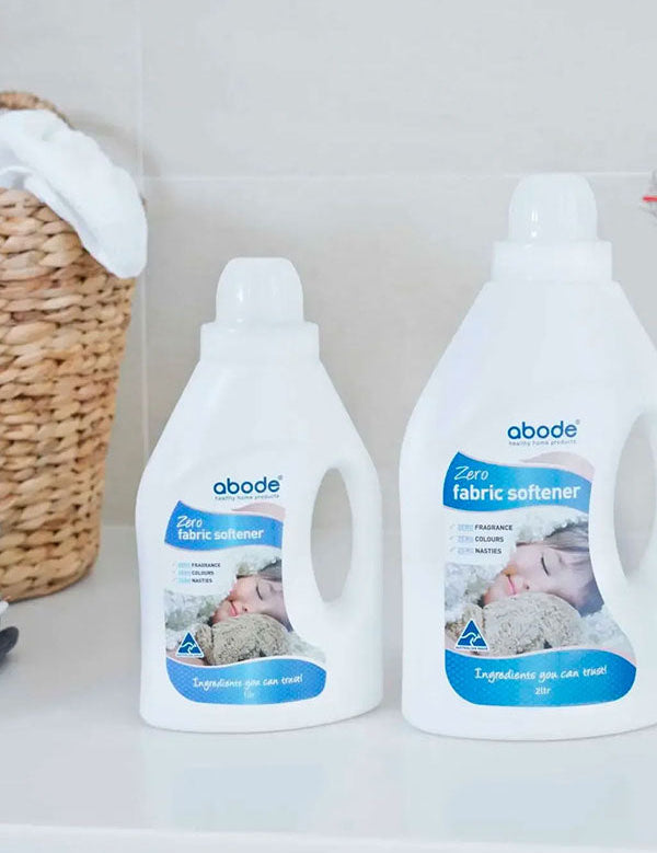 Two bottles of Abode Fabric Softener Zero on a white laundry bench beside a woven basket and folded towels