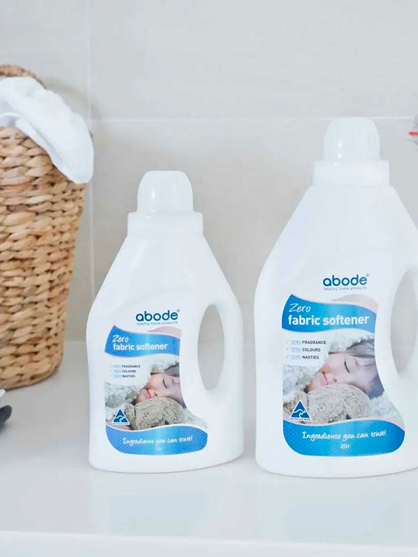 Two bottles of Abode Fabric Softener Zero on a white laundry bench beside a woven basket and folded towels