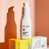 A white bottle of Acure Brightening Glowing Serum 30ml stands beside its yellow and white box on a peach background, both casting soft shadows and emphasizing the promise of radiant skin.