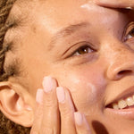 Model gently applying Acure Revived Radiance Overnight Mask to her face in warm sunlight.