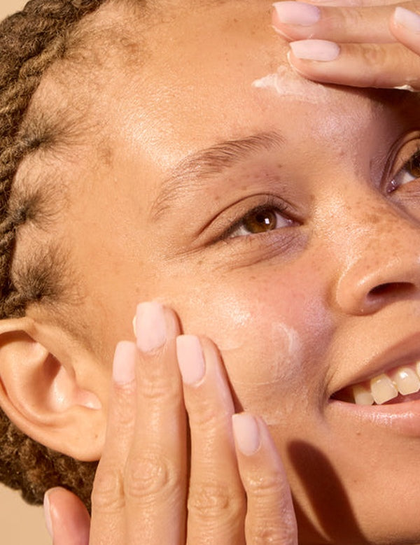 Model gently applying Acure Revived Radiance Overnight Mask to her face in warm sunlight.