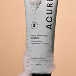 Acure Simply Smoothing Shampoo tube surrounded by rich white foam on a peach background.
