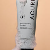 Acure Simply Smoothing Shampoo tube surrounded by rich white foam on a peach background.