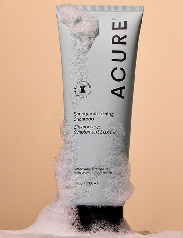 Acure Simply Smoothing Shampoo tube surrounded by rich white foam on a peach background.