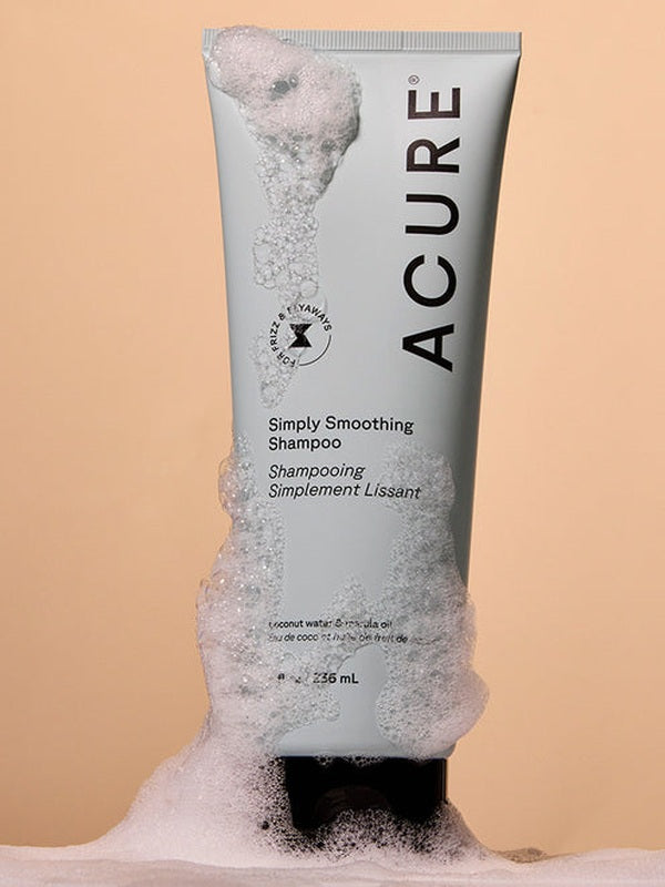 Acure Simply Smoothing Shampoo tube surrounded by rich white foam on a peach background.