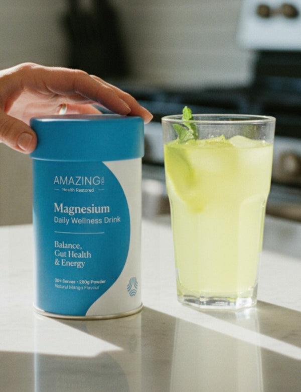 A hand rests on a blue Amazing You Daily Wellness Drink canister beside a glass of pale green iced drink garnished with mint.
