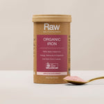 Amazonia Raw Organic Iron canister standing beside a gold spoon of pink powder on a pale tabletop against a beige background.