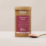 Amazonia Raw Organic Iron canister standing beside a gold spoon of pink powder on a pale tabletop against a beige background.