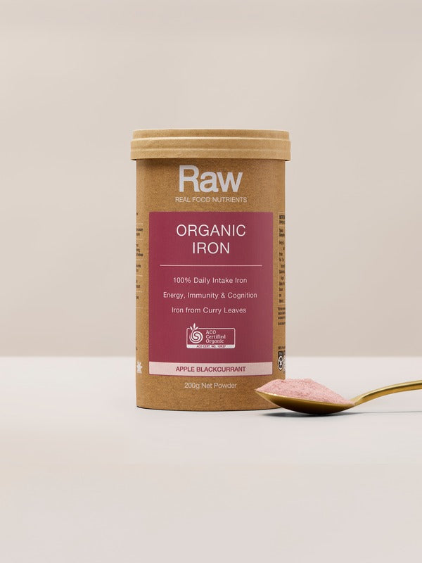 Amazonia Raw Organic Iron canister standing beside a gold spoon of pink powder on a pale tabletop against a beige background.