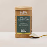 Amazonia Raw Organic Multivitamin canister standing beside a gold spoon filled with pale powder on a light tabletop against a beige background.