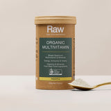 Amazonia Raw Organic Multivitamin canister standing beside a gold spoon filled with pale powder on a light tabletop against a beige background.