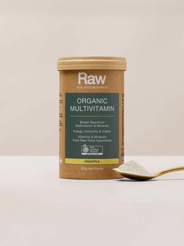 Amazonia Raw Organic Multivitamin canister standing beside a gold spoon filled with pale powder on a light tabletop against a beige background.