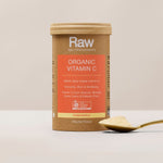 Amazonia Raw Organic Vitamin C canister standing beside a gold spoon heaped with pale powder on a light tabletop against a beige background.