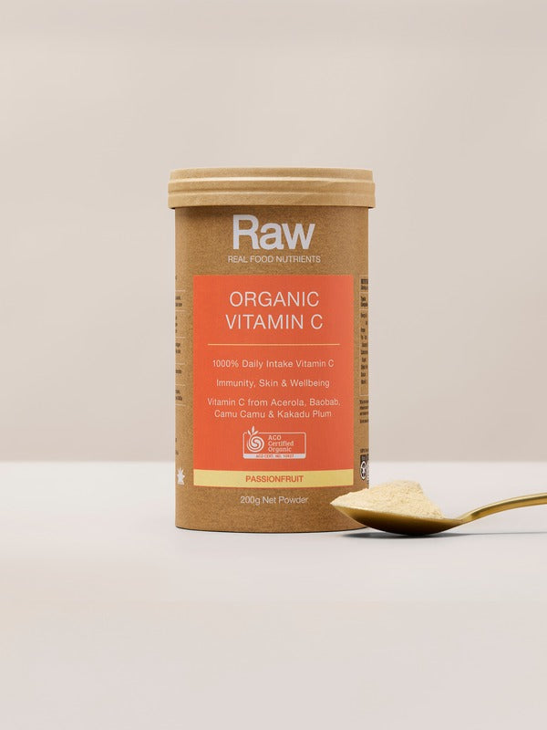 Amazonia Raw Organic Vitamin C canister standing beside a gold spoon heaped with pale powder on a light tabletop against a beige background.