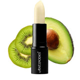Antipodes Kiwi Seed Oil Lip Conditioner 4g is pictured in front of a sliced kiwi and half an avocado, set on a white background.