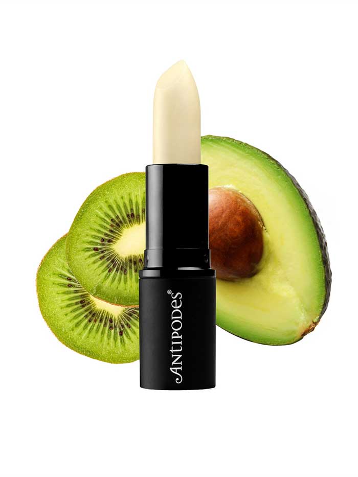 Antipodes Kiwi Seed Oil Lip Conditioner 4g is pictured in front of a sliced kiwi and half an avocado, set on a white background.