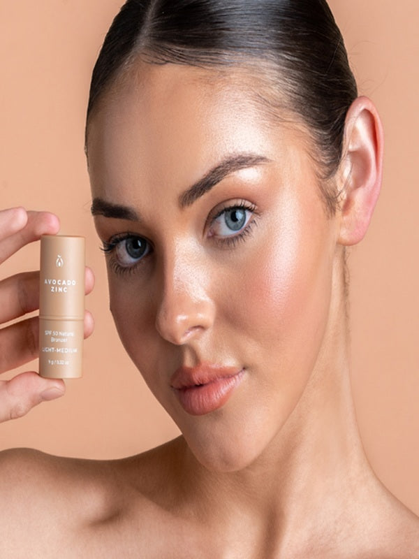 Model holding Avocado Zinc SPF50 Natural Bronzer in Light-Medium beside her highlighted cheek.