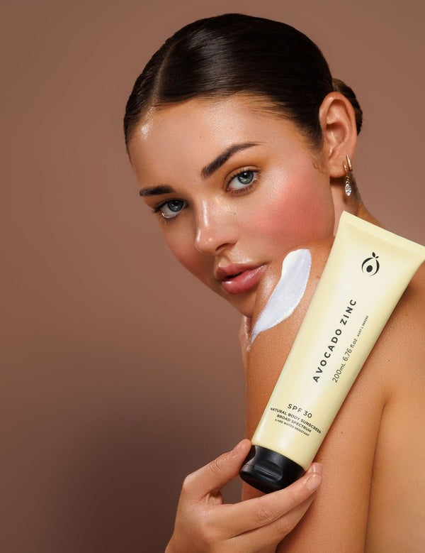 Model holding Avocado Zinc SPF30 Natural Body Sunscreen with a visible swipe of cream on her shoulder.