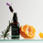 Amber Axilla deodorant spray bottle beside a purple lavender sprig and peeled orange, on a white surface edged in mint green.
