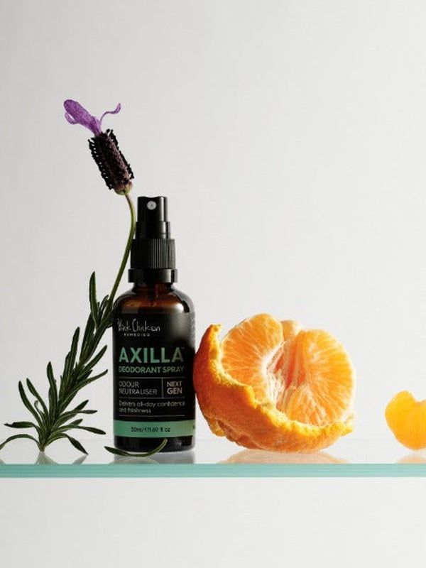 Amber Axilla deodorant spray bottle beside a purple lavender sprig and peeled orange, on a white surface edged in mint green.
