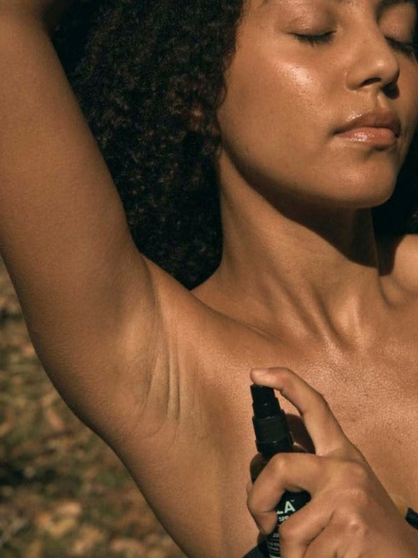 A woman with dark curly hair raises one arm and holds a black-pump Axilla deodorant bottle beneath her underarm, with her eyes closed in warm sunlight.