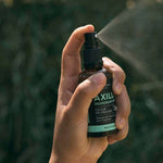 A hand presses the black pump of an amber Axilla deodorant bottle, releasing a fine mist to the right against blurred outdoor greenery.