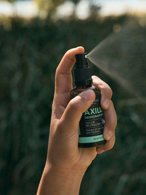 A hand presses the black pump of an amber Axilla deodorant bottle, releasing a fine mist to the right against blurred outdoor greenery.