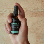 A hand holds an amber Axilla deodorant spray bottle upright, with a finger on the black pump and its turquoise lettering facing forward against a pale tan wall.