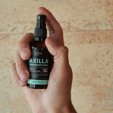 A hand holds an amber Axilla deodorant spray bottle upright, with a finger on the black pump and its turquoise lettering facing forward against a pale tan wall.
