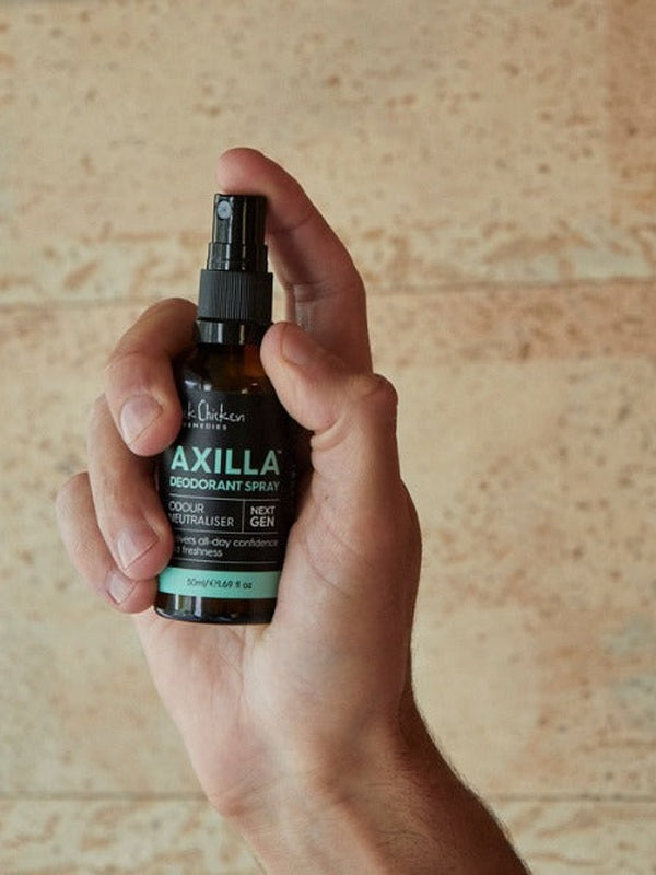 A hand holds an amber Axilla deodorant spray bottle upright, with a finger on the black pump and its turquoise lettering facing forward against a pale tan wall.