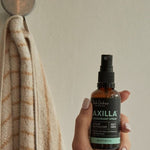 A hand holds an amber Axilla deodorant spray bottle beside a beige towel hanging from a round wall hook.
