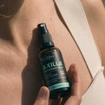 Close-up of a hand holding an amber Axilla deodorant spray bottle against a person's upper chest, beside the strap of a beige top.