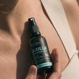 Close-up of a hand holding an amber Axilla deodorant spray bottle against a person's upper chest, beside the strap of a beige top.