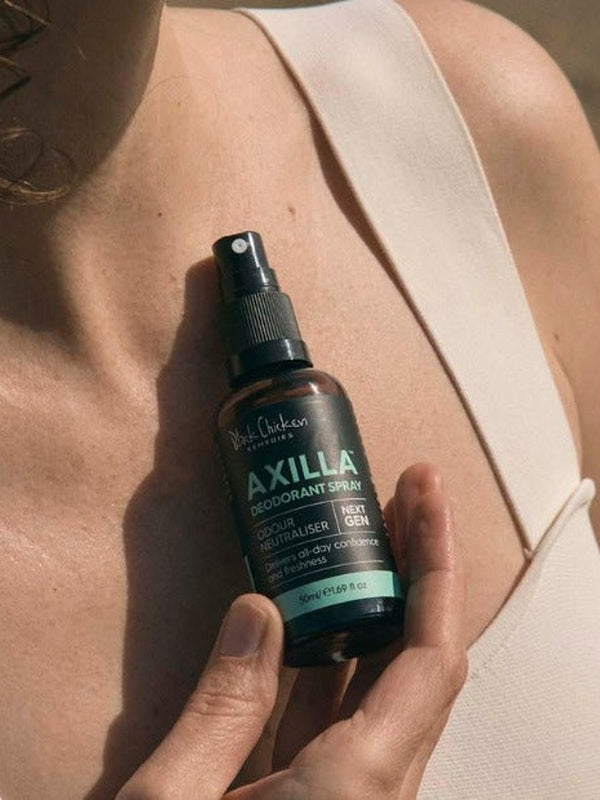 Close-up of a hand holding an amber Axilla deodorant spray bottle against a person's upper chest, beside the strap of a beige top.