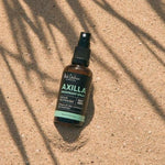 An amber Axilla deodorant spray bottle lies diagonally on sand beneath long, narrow leaf shadows.