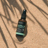 An amber Axilla deodorant spray bottle lies diagonally on sand beneath long, narrow leaf shadows.