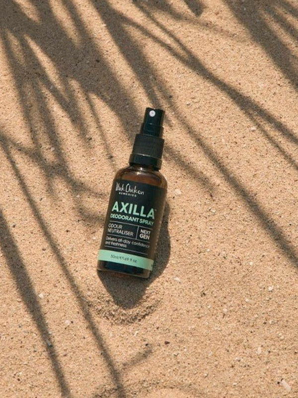 An amber Axilla deodorant spray bottle lies diagonally on sand beneath long, narrow leaf shadows.