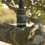 An amber Axilla deodorant spray bottle stands upright on a thick tree branch, surrounded by leaves and dappled sunlight.
