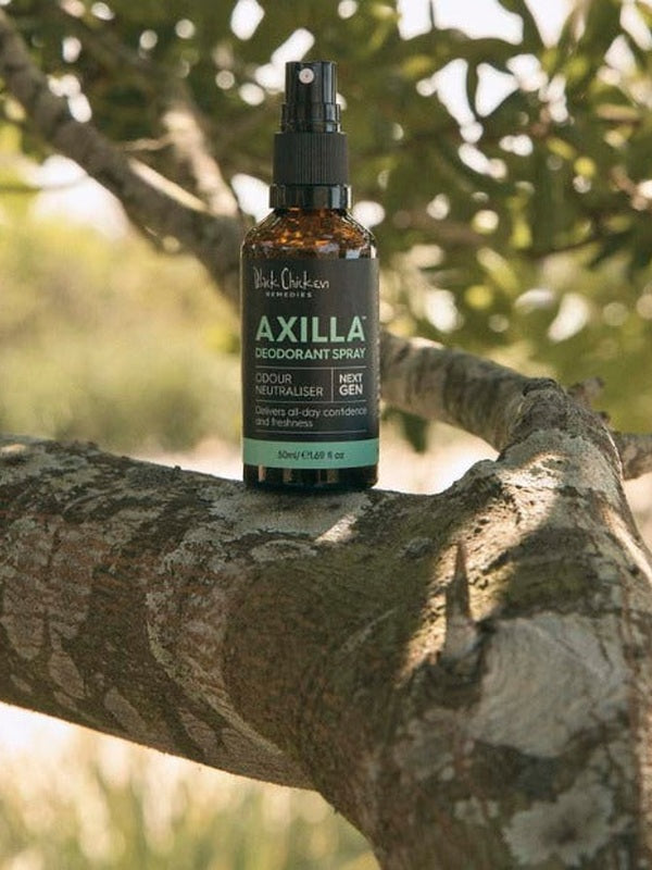 An amber Axilla deodorant spray bottle stands upright on a thick tree branch, surrounded by leaves and dappled sunlight.