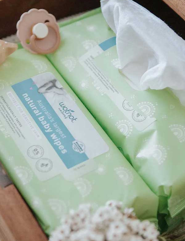 Two packs of Wotnot Natural Baby Wipes 70pk in green packaging, one partly open with a sheet out, are displayed in a wooden box beside a baby pacifier and white flowers.